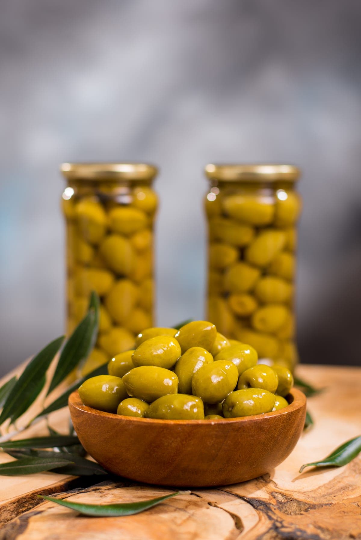 Jars of green olives next to a wooden bowl with an olive branch
